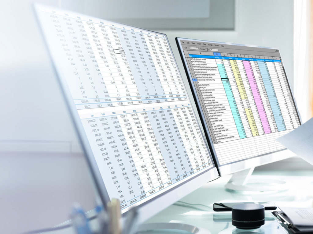 Two monitors on a desk, both displaying excel spreadsheets