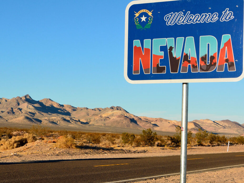 Welcome to Nevada sign on the side of the roadway passing through an arid stretch of land.